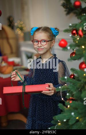 Ein fröhliches kleines Mädchen in stilvoller Brille und einem wunderschönen festlichen Kleid steht neben einem glitzernden Weihnachtsbaum und legt ein wunderschön gewickeltes Geschenk in ihre Hände. Moment, um ihr Geschenk auszupacken, das den zauberhaften Geist des Weihnachtsstils verkörpert. Stockfoto