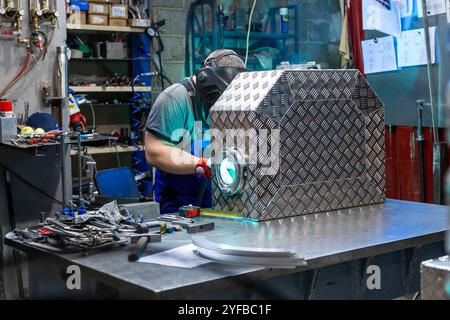 Ein Arbeiter, der Schutzausrüstung trägt, schweißt in einer gut ausgestatteten Werkstatt einen Metallkasten und betont dabei Präzision und Handwerkskunst. Stockfoto