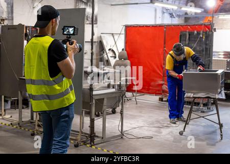 Ein Mann in einer gelben Weste filmt einen Arbeiter, der Metallgeräte in einer Industriewerkstatt bearbeitet und den Herstellungsprozess dokumentiert. Stockfoto