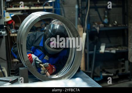 Schweißer arbeitet geschickt an einem großen Metallring, schweisst sorgfältig Verbindungen in einer gut ausgestatteten Werkstatt. Stockfoto