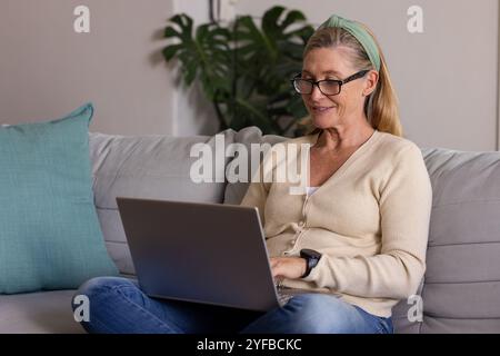 Lächelnde ältere Frau, die zu Hause einen Laptop auf einem gemütlichen Sofa benutzt Stockfoto