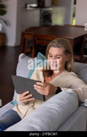 Ältere Frau, die sich zu Hause mit Tablet auf dem Sofa entspannt und zufrieden lächelt Stockfoto