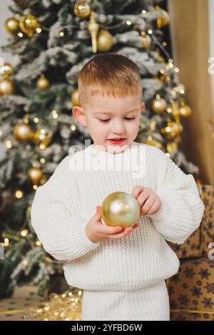 Ein fröhlicher kleiner Junge von 2-3 Jahren steht neben einem Weihnachtsbaum, umgeben von Girlanden und Weihnachtsgeschenken. Das Konzept eines Weihnachtstals Stockfoto