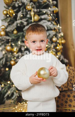 Ein fröhlicher kleiner Junge von 2-3 Jahren steht neben einem Weihnachtsbaum, umgeben von Girlanden und Weihnachtsgeschenken. Das Konzept eines Weihnachtstals Stockfoto