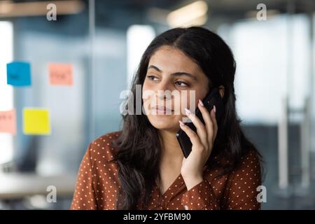Junge hispanische Geschäftsfrau in moderner Büroumgebung am Telefon, die Fokus ausdrückt. Professionelle Kleidung und Haftnotizen im Hintergrund deuten auf eine kreative Atmosphäre am Arbeitsplatz hin. Stockfoto