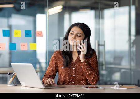 Junge hispanische Geschäftsfrau Multitasking mit Telefon und Laptop im Büro. Professional konzentriert sich während der Arbeit am Computer auf Anrufe und repräsentiert Geschäftskommunikation, Technologie und Produktivität. Stockfoto