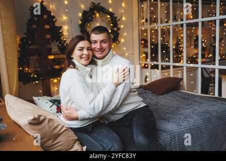 Familienpaar in warmen Strickpullis auf dem Bett mit Weihnachtsdekoration. Neujahrsdekoration in einem gemütlichen Haus. Stockfoto