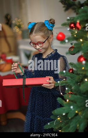 Ein fröhliches kleines Mädchen in stilvoller Brille und einem wunderschönen festlichen Kleid steht neben einem glitzernden Weihnachtsbaum und legt ein wunderschön gewickeltes Geschenk in ihre Hände. Moment, um ihr Geschenk auszupacken, das den zauberhaften Geist des Weihnachtsstils verkörpert. Stockfoto