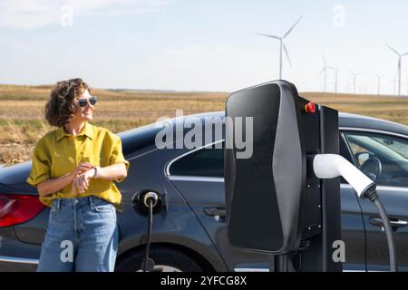 Eine Frau mit gewölbten Haaren, die ein gelbes Hemd und eine Sonnenbrille trägt, steht neben dem Aufladen des Elektroautos. Windräder im Hintergrund. Stockfoto