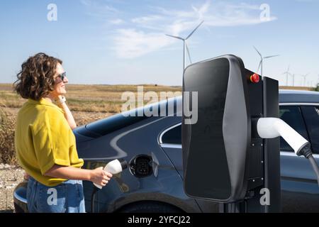 Eine Frau mit gewölbten Haaren, die ein gelbes Hemd und eine Sonnenbrille trägt, steht neben dem Aufladen des Elektroautos. Windkraftanlage im Hintergrund. Stockfoto