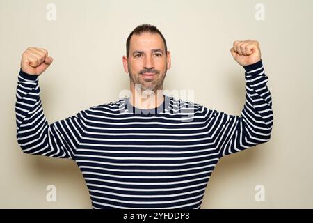 Bärtiger hispanischer Mann in seinen 40ern, der einen gestreiften Pullover trug, seinen Bizeps beugte, während er stolz lächelte, ohne die bedauerliche körperliche Verfassung zu bemerken Stockfoto
