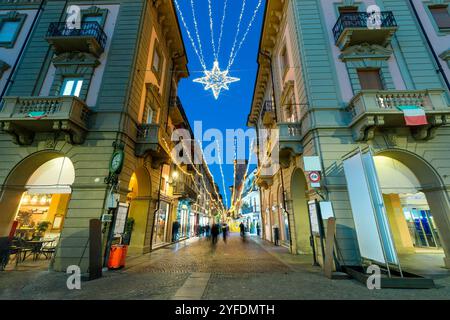 Fußgängerzone mit Kopfsteinpflaster zwischen Gebäuden, beleuchtet und mit Weihnachtslichtern dekoriert in Alba, Italien. Stockfoto