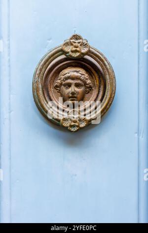 Ein antiker Türklopfer aus Messing mit klassischem Gesicht mit Blumenelementen, der vor einer hellblauen Holztür steht. Dieses Bild weckt einen Sinn für Histoismus Stockfoto
