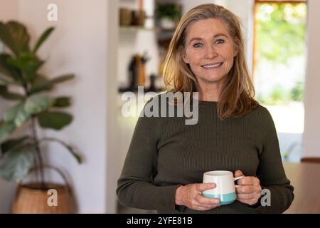 Lächelnde Seniorin hält zu Hause eine Kaffeetasse und genießt einen gemütlichen Morgen Stockfoto