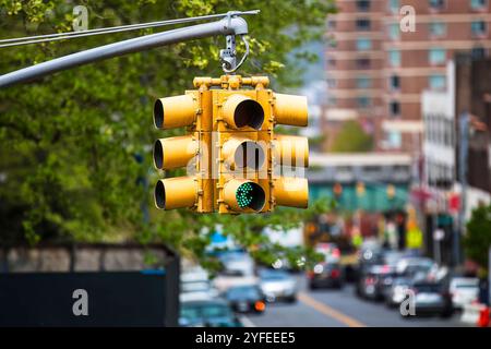 Eine gelbe Ampel in New York City hängt an einem gebogenen Metallmast und zeigt ein grünes Signal mit einem verschwommenen Hintergrund von Autos und Gebäuden. Stockfoto