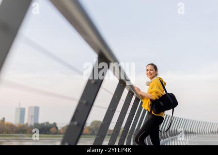 Eine junge Frau in einer leuchtend gelben Jacke lehnt sich an ein modernes Geländer, während sie sich auf ihr Handy konzentriert und einen ruhigen Spaziergang am Fluss auf einem Pett genießt Stockfoto