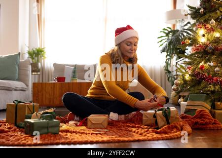 Seniorin, die Geschenke am Weihnachtsbaum verpackt, festliche Feiertagsstimmung genießt, zu Hause Stockfoto