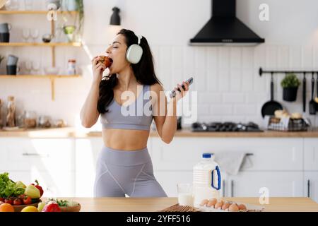 Die junge Sportlerin genießt einen gesunden Apfel-Snack, während sie in ihrer Küche Musik auf ihrem Smartphone hört Stockfoto