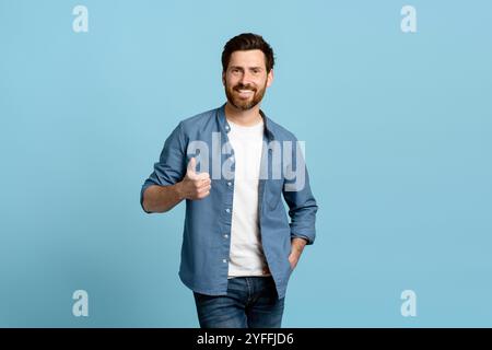 Selbstbewusster junger Mann vor blauem Hintergrund lächelt und zeigt einen Daumen hoch, strahlt Positivität und Erfolg aus. Er trägt ein weißes Hemd, ein blaues Hemd, Stockfoto