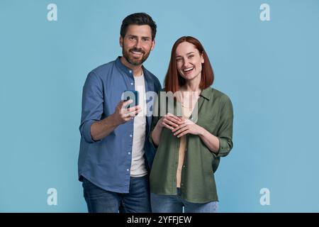 Fröhlicher junger erwachsener Mann und Frau lachen, während sie zusammen ein Smartphone benutzen, vor blauem Hintergrund Stockfoto