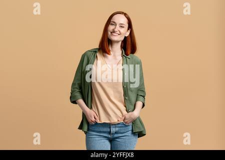 Eine junge Frau mit rotem Haar steht mit den Händen in den Taschen und lächelt auf beigefarbenem Hintergrund Stockfoto