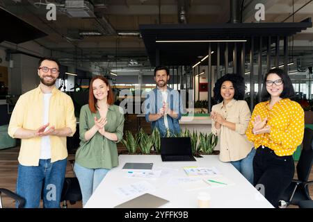 Eine Gruppe von fünf verschiedenen Geschäftsleuten applaudiert, während sie an einem Tisch in einem modernen Büro stehen Stockfoto