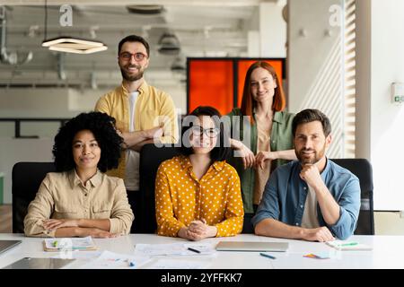 Ein vielseitiges Team von fünf Geschäftsleuten lächelt, während sie in einem modernen Büro um einen Tisch sitzen und stehen Stockfoto