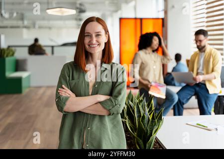 Porträt einer lächelnden Geschäftsfrau, die mit überkreuzten Armen in einem modernen Büro steht, während ihr Team im Hintergrund arbeitet Stockfoto