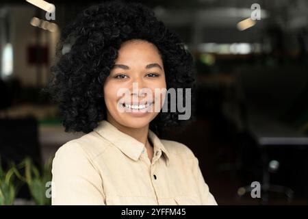 Eine junge Geschäftsfrau mit lockigem Haar lächelt in einer Büroumgebung selbstbewusst Stockfoto
