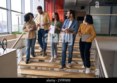 Ein vielseitiges Team von Fachleuten läuft in einem Bürogebäude die Treppe hinunter, führt Gespräche und trägt Laptops und Kaffee mit sich Stockfoto