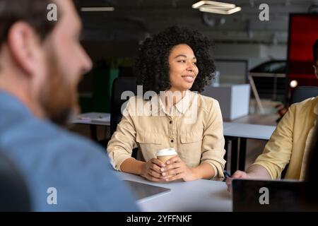 Die junge Führungskraft hält eine Kaffeetasse in der Hand und hört den Kollegen während eines Firmensitzes im Büro zu Stockfoto