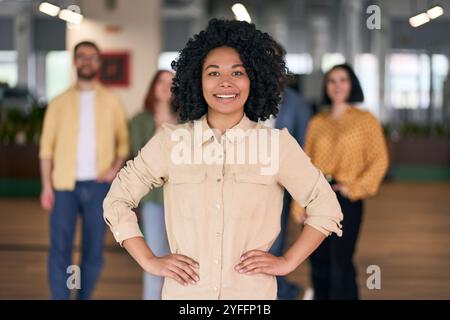 Die junge, selbstbewusste Geschäftsfrau posiert mit den Händen auf den Hüften, während ihr Team hinter ihr steht Stockfoto
