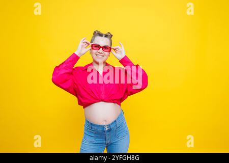 Glückliche Schwangerschaft. Fröhliche, hübsche schwangere Frau in einem rosa Hemd und einer rosa Brille auf gelbem Hintergrund. Junge helle schwangere Frau. Stockfoto