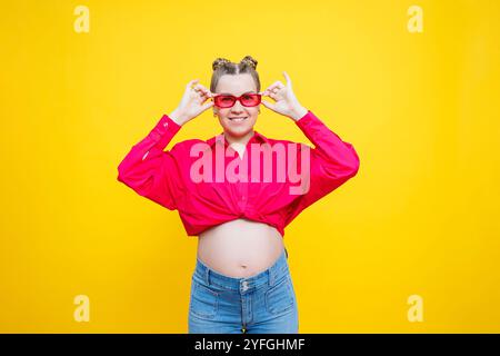 Glückliche Schwangerschaft. Fröhliche, hübsche schwangere Frau in einem rosa Hemd und einer rosa Brille auf gelbem Hintergrund. Junge helle schwangere Frau. Stockfoto