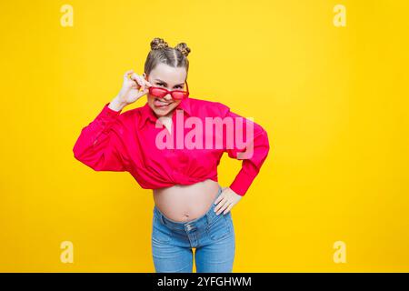 Glückliche Schwangerschaft. Fröhliche, hübsche schwangere Frau in einem rosa Hemd und einer rosa Brille auf gelbem Hintergrund. Junge helle schwangere Frau. Stockfoto