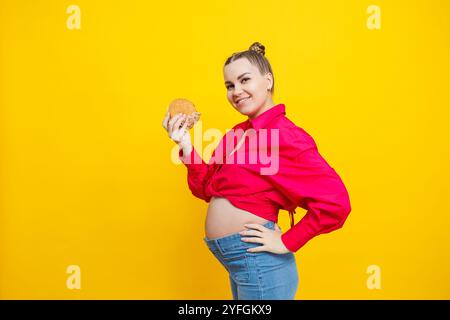 Lustige schwangere junge Frau, die Burger isst. Schwangere Frau, die leckeres Sandwich hält, während sie isoliert auf der gelben Studiowand steht. Junk Food Konzept. Schnell Stockfoto