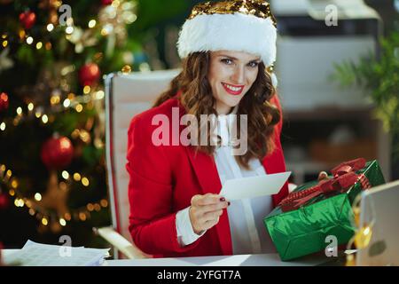 Weihnachten. Porträt einer glücklichen, stilvollen Geschäftsfrau mit weihnachtsmütze und roter Jacke mit Geschenkbox und Postkarte im modernen grünen Büro mit Christm Stockfoto