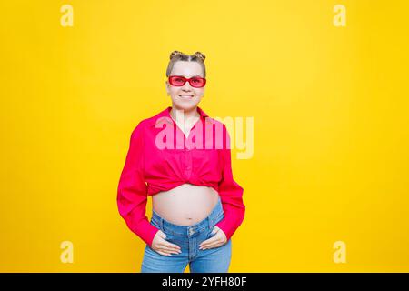 Glückliche Schwangerschaft. Fröhliche, hübsche schwangere Frau in einem rosa Hemd und einer rosa Brille auf gelbem Hintergrund. Junge helle schwangere Frau. Stockfoto