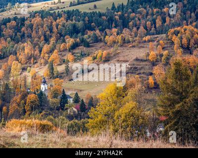 Idyllische Herbstlandschaft mit einer malerischen Kirche, umgeben von bunten Blättern und einem Hirten, der auf einem Hügel Schafherde pflegt Stockfoto