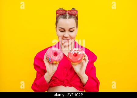 Lächelnde schwangere Frau mit süßen Kuchen in den Händen auf gelbem Hintergrund. Köstliche süße Donuts in den Händen einer Frau. Schädliche Lebensmittel Stockfoto