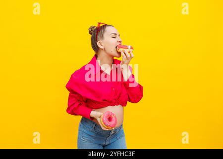 Lächelnde schwangere Frau mit süßen Kuchen in den Händen auf gelbem Hintergrund. Köstliche süße Donuts in den Händen einer Frau. Schädliche Lebensmittel Stockfoto