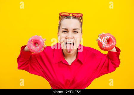 Lächelnde schwangere Frau mit süßen Kuchen in den Händen auf gelbem Hintergrund. Köstliche süße Donuts in den Händen einer Frau. Schädliche Lebensmittel Stockfoto