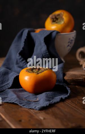 Reife Persimmon sitzt auf einem dunkelblauen Tuch, mit einer Keramikschale im Hintergrund und schafft ein rustikales, herbstliches Stillleben auf einem Holztisch Stockfoto