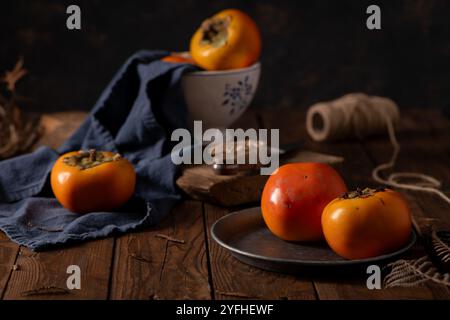 Persimmonen ruhen auf einem rustikalen Holztisch, umgeben von einer Keramikschale, einem dunkelblauen Tuch, Garn und natürlichen Elementen, die herbstliche Wärme hervorrufen Stockfoto