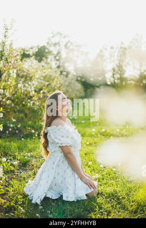 Eine glückliche schwangere Frau in einem sommerweißen Musselin-Kleid sitzt auf dem Gras in einem blühenden Garten. Zukünftige Mutter, eine Frau, die ein Baby erwartet. Schwangerschaft Stockfoto