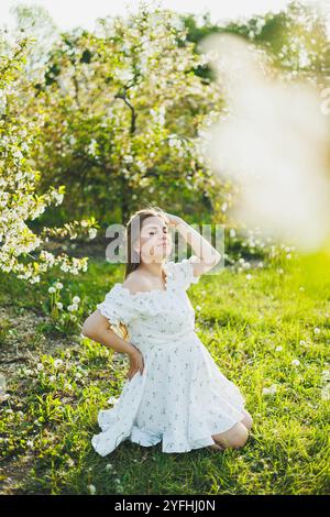 Eine glückliche schwangere Frau in einem sommerweißen Musselin-Kleid sitzt auf dem Gras in einem blühenden Garten. Zukünftige Mutter, eine Frau, die ein Baby erwartet. Schwangerschaft Stockfoto