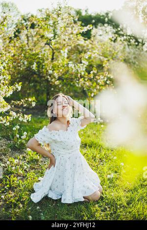 Eine glückliche schwangere Frau in einem sommerweißen Musselin-Kleid sitzt auf dem Gras in einem blühenden Garten. Zukünftige Mutter, eine Frau, die ein Baby erwartet. Schwangerschaft Stockfoto