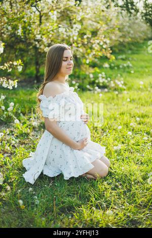 Eine glückliche schwangere Frau in einem sommerweißen Musselin-Kleid sitzt auf dem Gras in einem blühenden Garten. Zukünftige Mutter, eine Frau, die ein Baby erwartet. Schwangerschaft Stockfoto