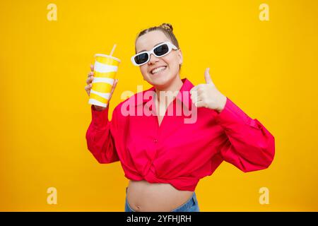 Eine süße schwangere Frau in einem rosa Hemd trinkt Wasser aus einem Plastikbecher. Eine schwangere Frau auf gelbem Hintergrund mit einem Glas in der Hand. Schädliches Wasser Stockfoto