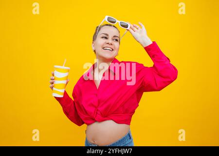 Eine süße schwangere Frau in einem rosa Hemd trinkt Wasser aus einem Plastikbecher. Eine schwangere Frau auf gelbem Hintergrund mit einem Glas in der Hand. Schädliches Wasser Stockfoto